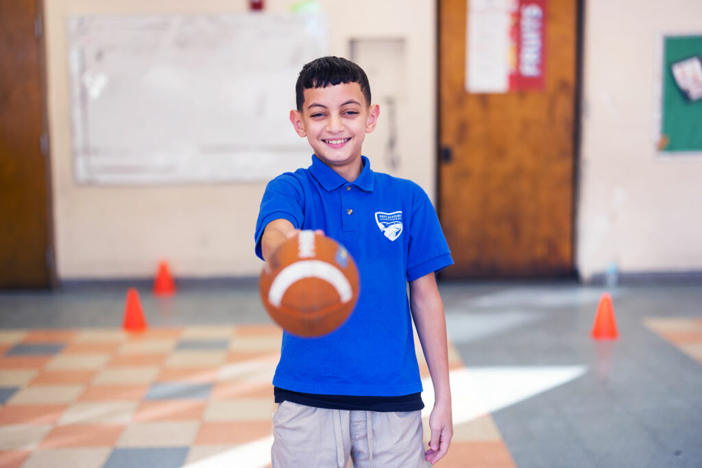 Smiling student wearing a Northwest Academy polo holding a football in a classroom.